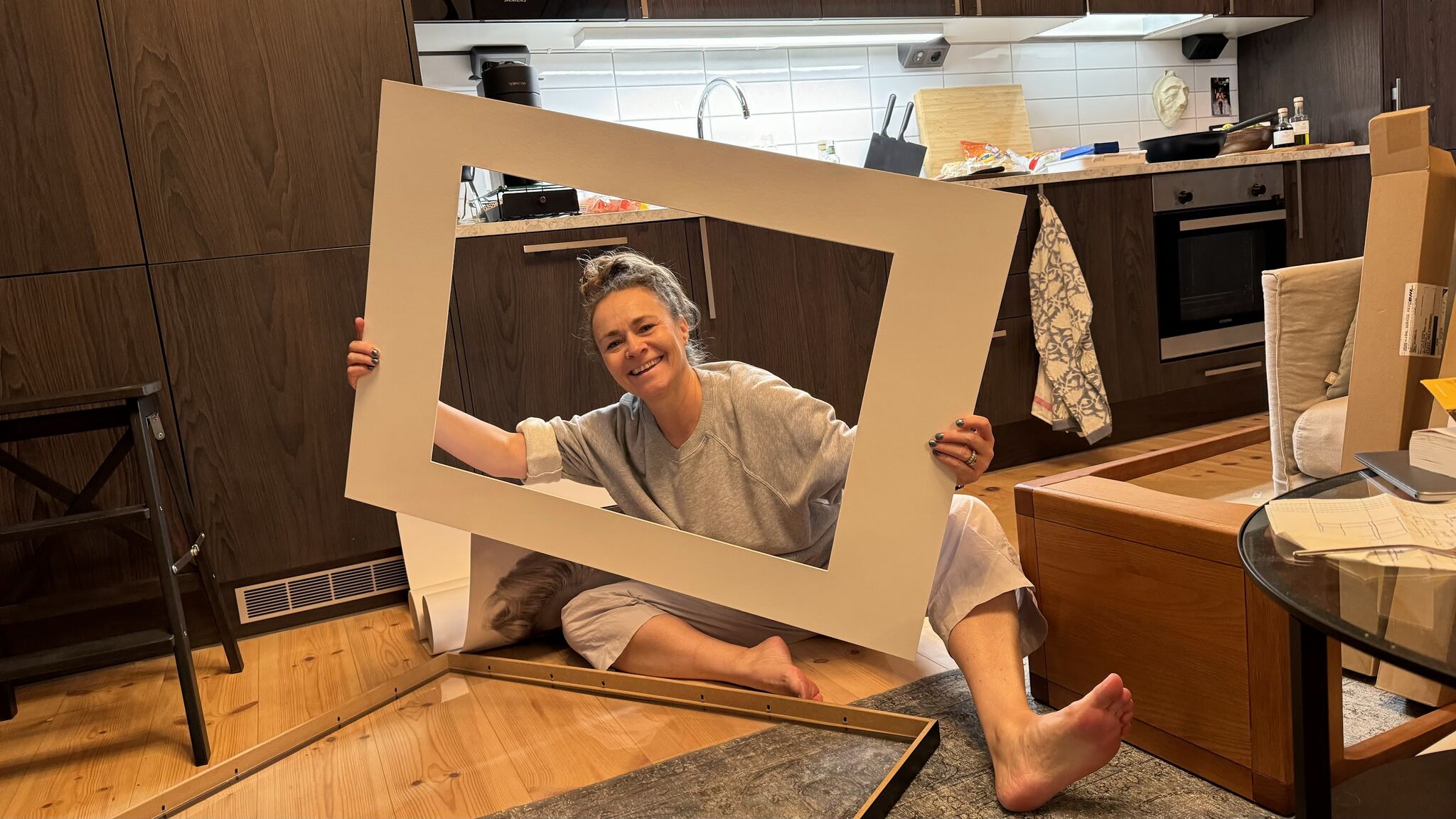 A smiling woman holds a large empty frame while sitting among furniture pieces in a kitchen.